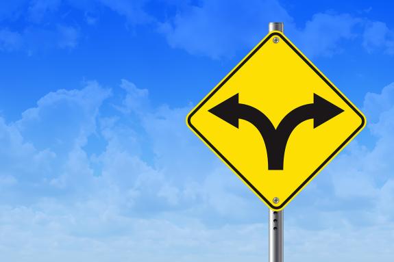 A yellow road sign with a diverging arrow symbolises a critical crossroads against a blue sky.